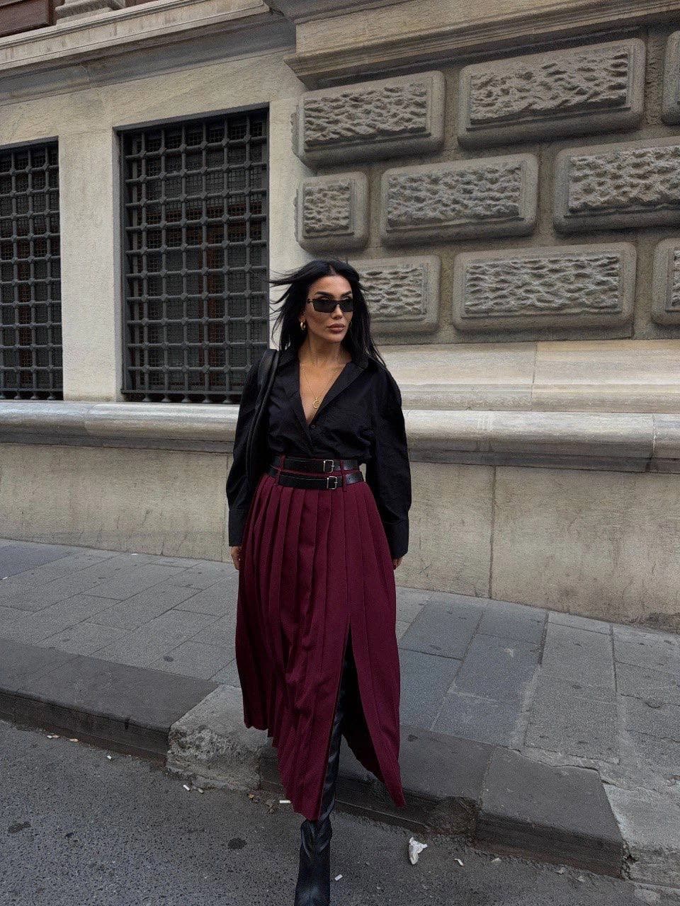 Burgundy pleated midi skirt featuring a high slit, cinched with a double belt harness. Elegant and modern fashion.