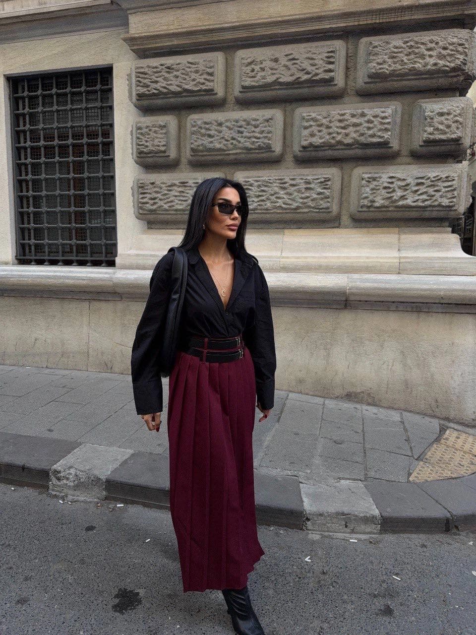 Burgundy midi skirt, neatly accordion-folded, featuring a dual-strap waist harness. Model presents a refined modern l...