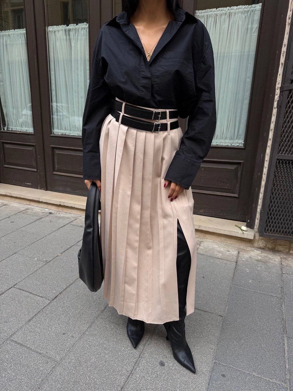 Cream high-waisted pleated midi skirt with a side slit and black double buckle belt, worn with a black shirt.